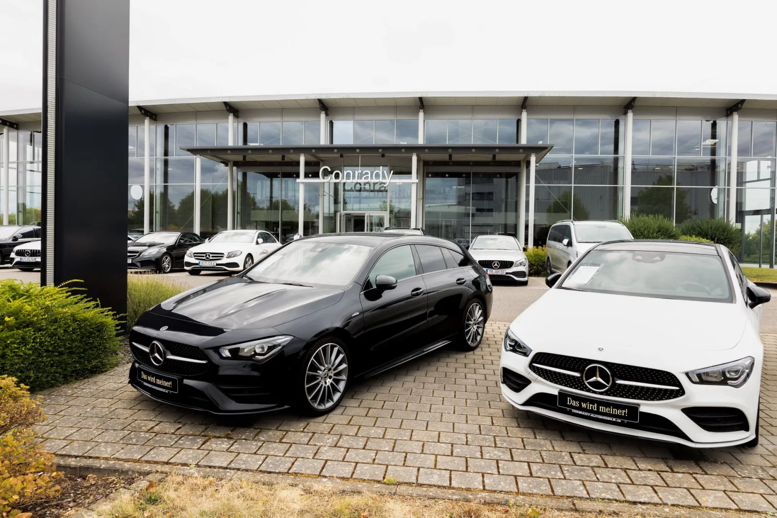 Ein schwarzer und ein weißer Mercedes CLA Shooting Brake vor dem Eingang von Conrady Automobile