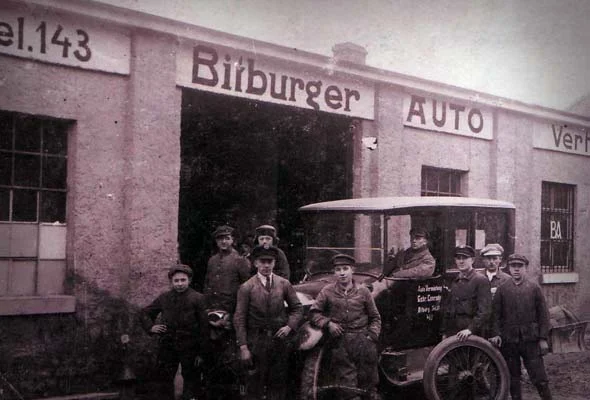 Belegschaft mit Schiebermützen vor der Halle des Bitburger Auto-Vertriebs, daneben ein frühes Automobil